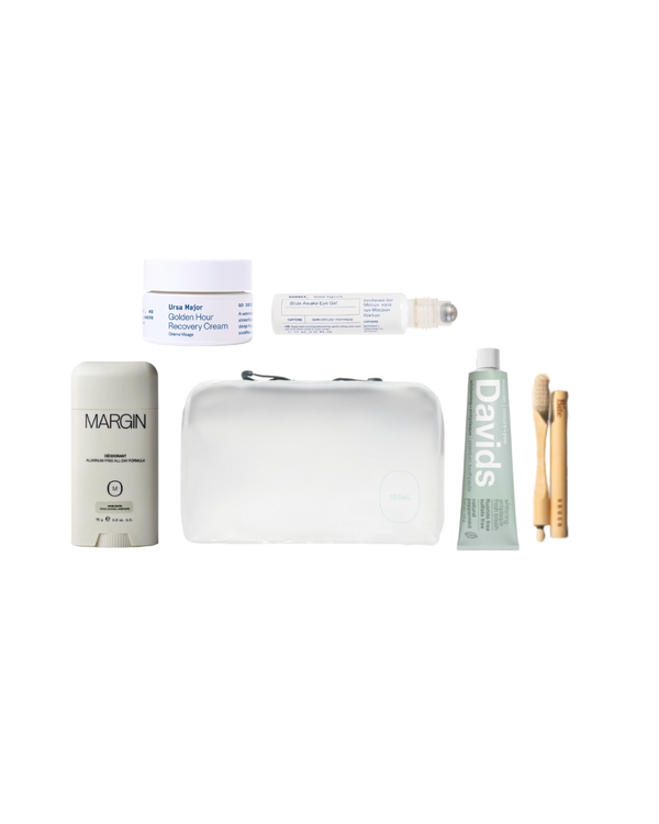 A curated travel self-care gift set arranged on a white background, including a Margin deodorant stick, a white travel cream jar, a clear roll-on bottle, a white zip pouch, a green Davids toothpaste tube, two bamboo toothbrushes, and a small white bottle.